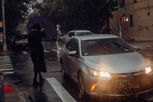 A person with an umbrella crosses a wet urban street in the rain, near a silver car.