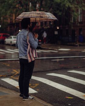 A man stands on a wet city crosswalk holding an umbrella during a rainy day.