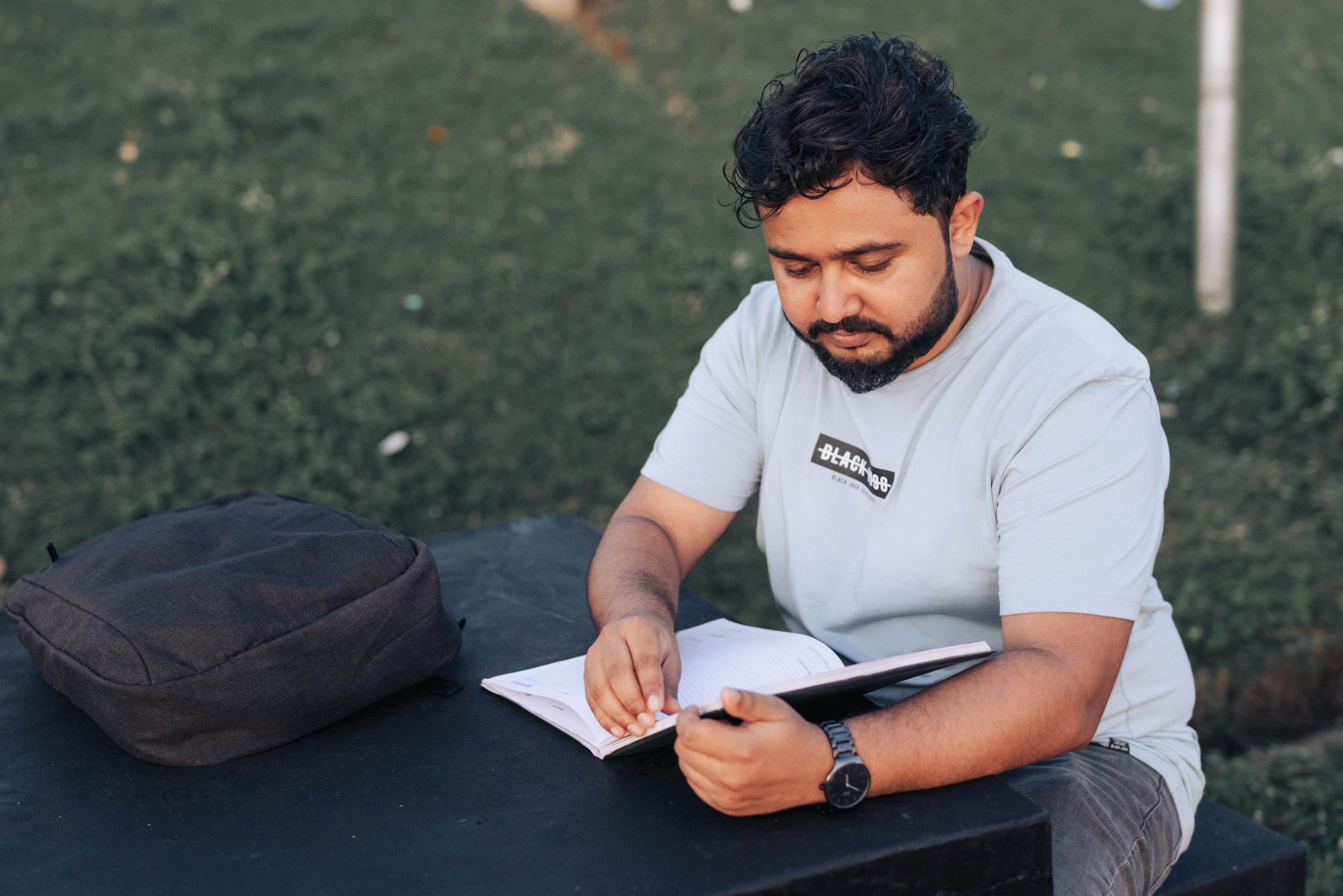 A young man in casual attire studying outdoors with a notebook and backpack.
