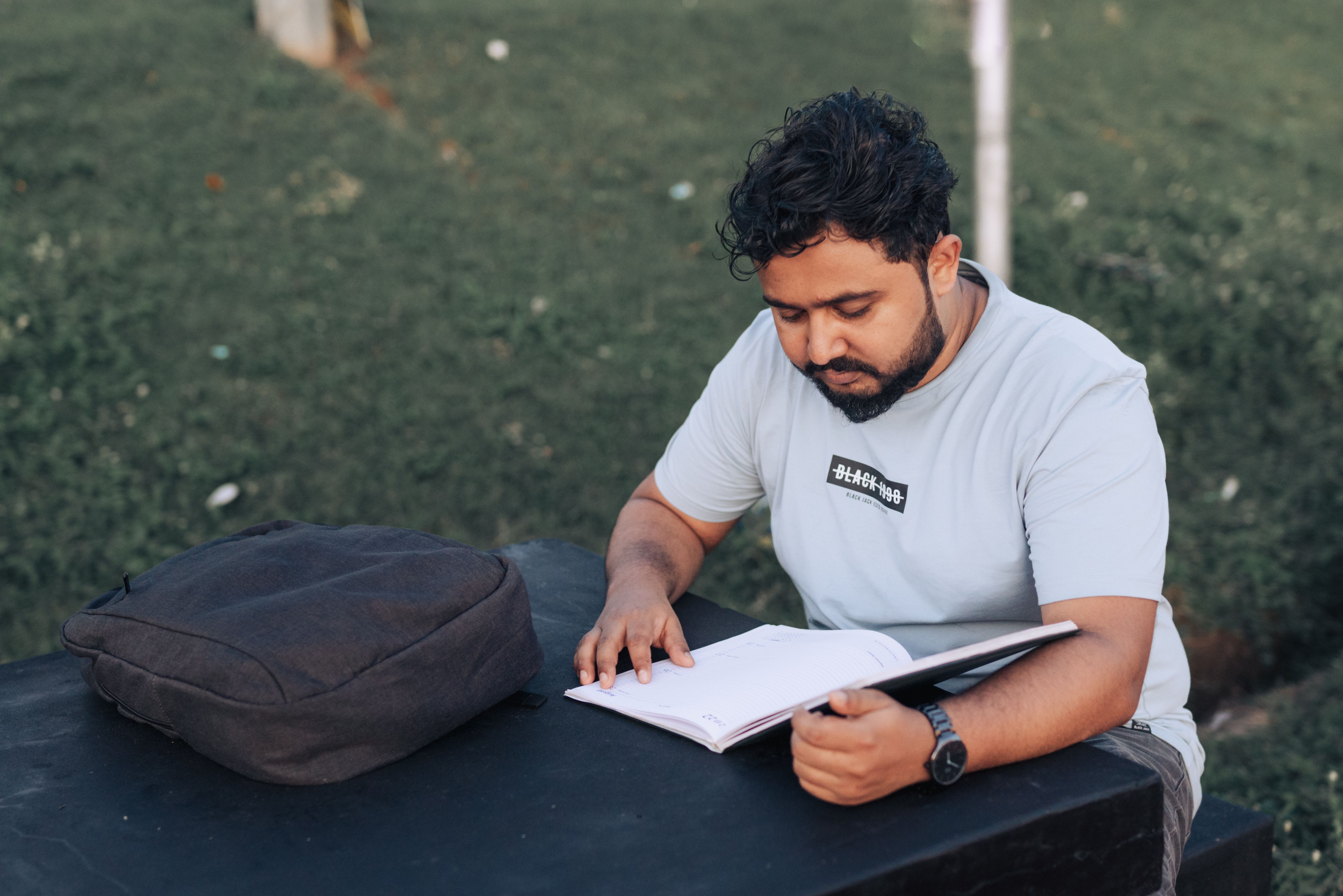A man with a beard reading a notebook outdoors on a sunny day, wearing a t-shirt.