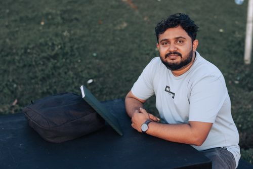 Free A young man sits outdoors at a table with a laptop and backpack, enjoying a serene park setting. Stock Photo