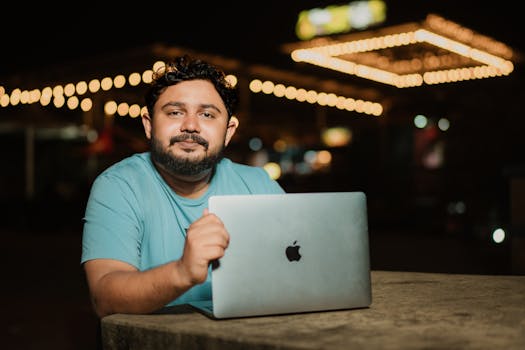 A man in a t-shirt sits outside using a laptop at night with string lights in the background.