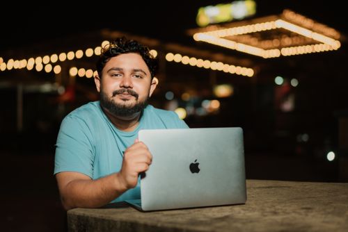 Free A man in a t-shirt sits outside using a laptop at night with string lights in the background. Stock Photo