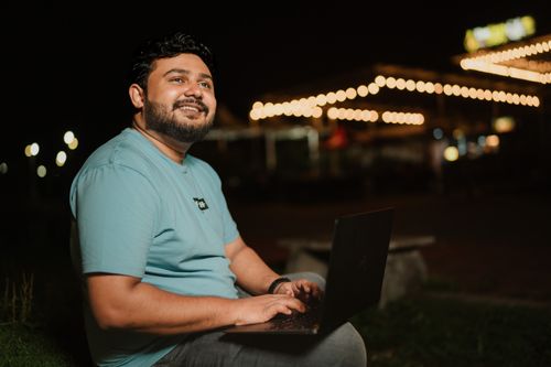 Free Smiling man typing on laptop outdoors at night with string lights in the background. Stock Photo