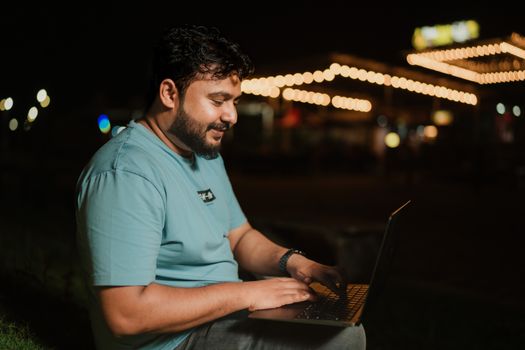 A man in a blue t-shirt typing on a laptop outdoors under string lights at night.