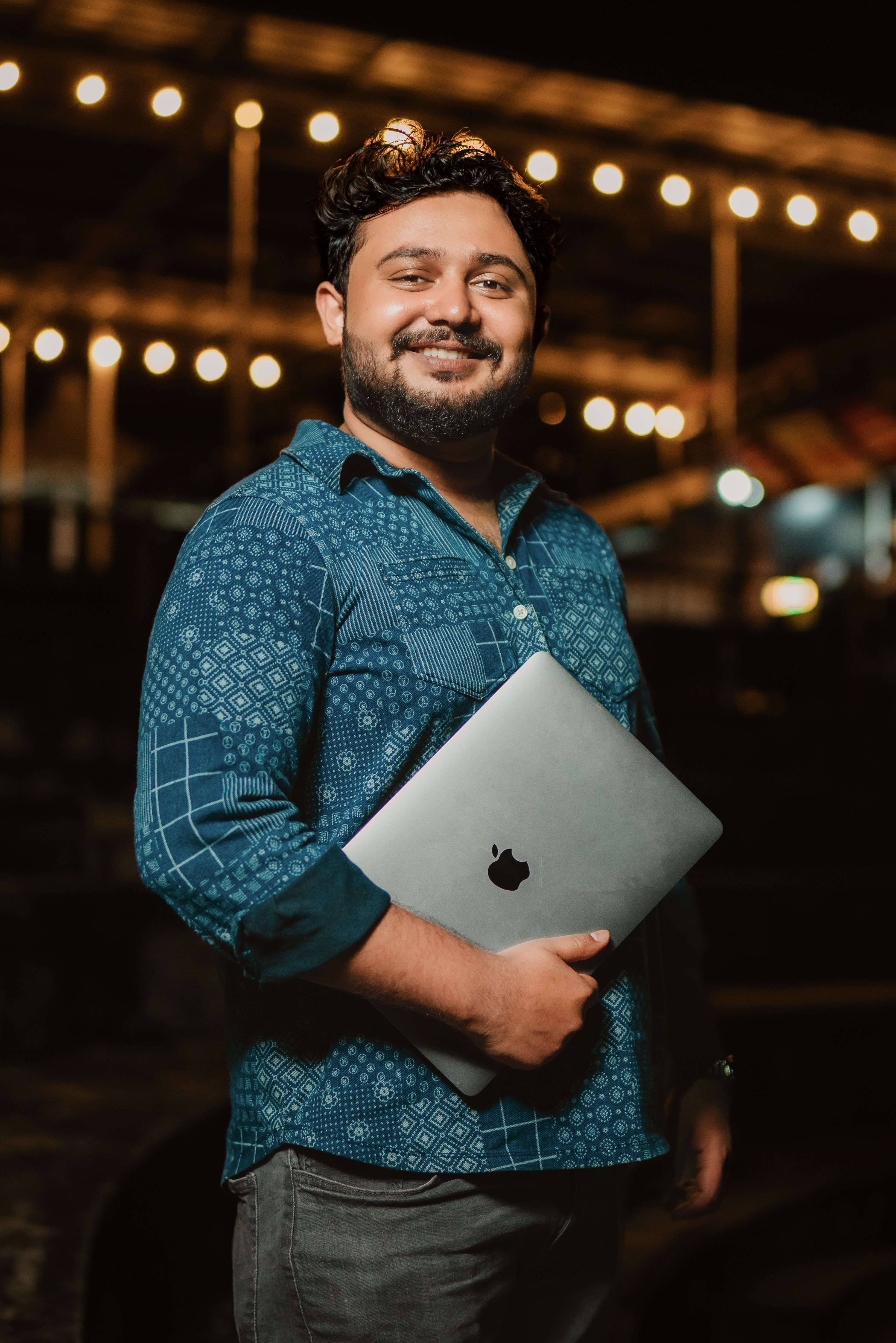 A happy man with a beard holding a laptop outdoors at night, surrounded by bokeh lights.