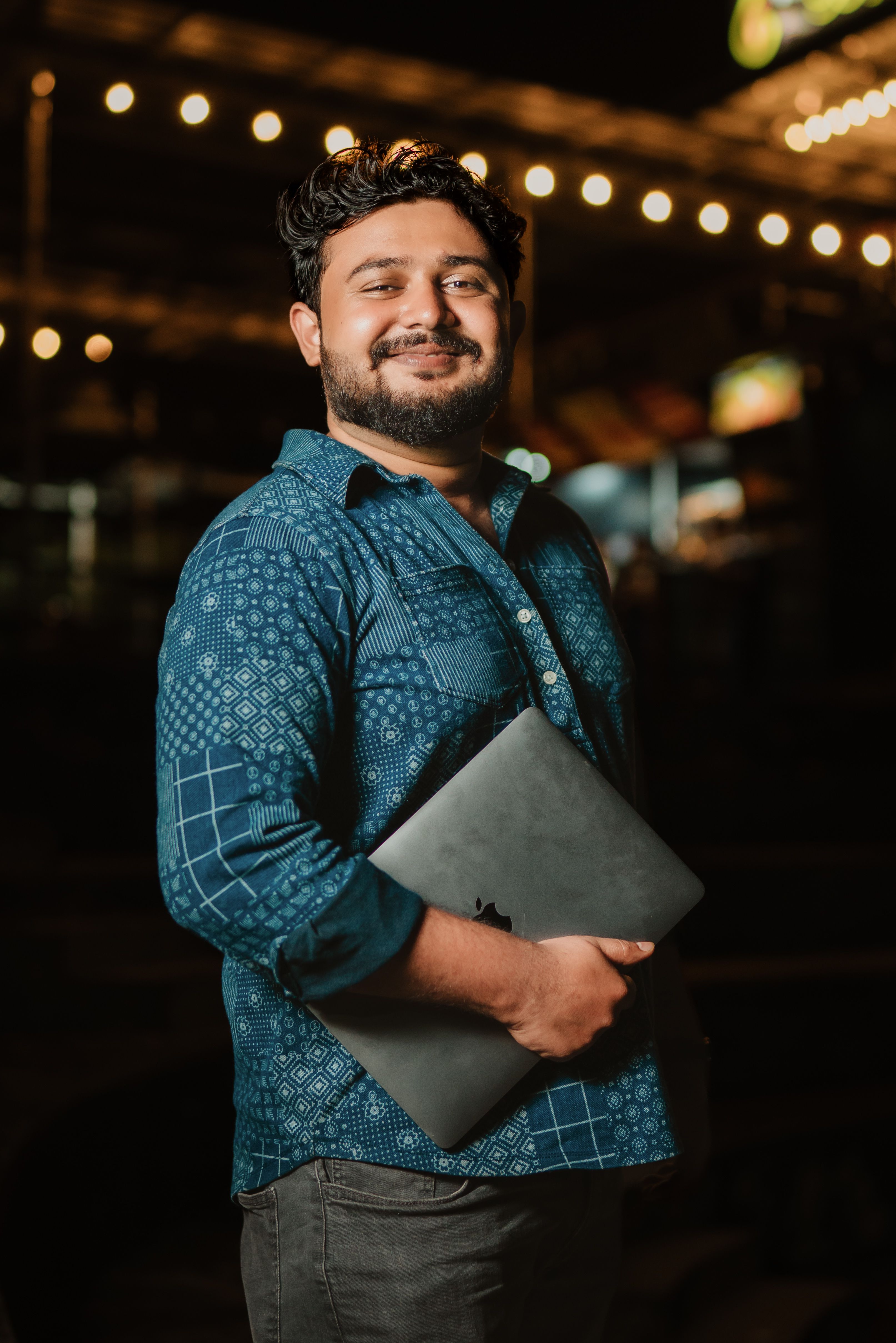 A happy man holding a laptop smiles confidently at night with decorative lights behind him.