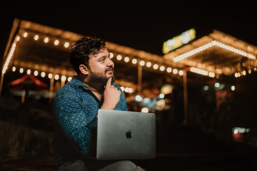 A thoughtful adult man with a beard using a laptop at night in a brightly lit outdoor setting.