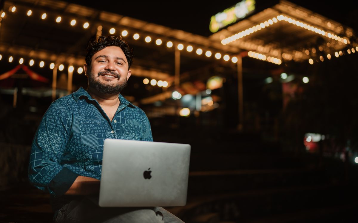 A cheerful adult man with a laptop, enjoying the night ambiance of a beautifully lit outdoor cafe.