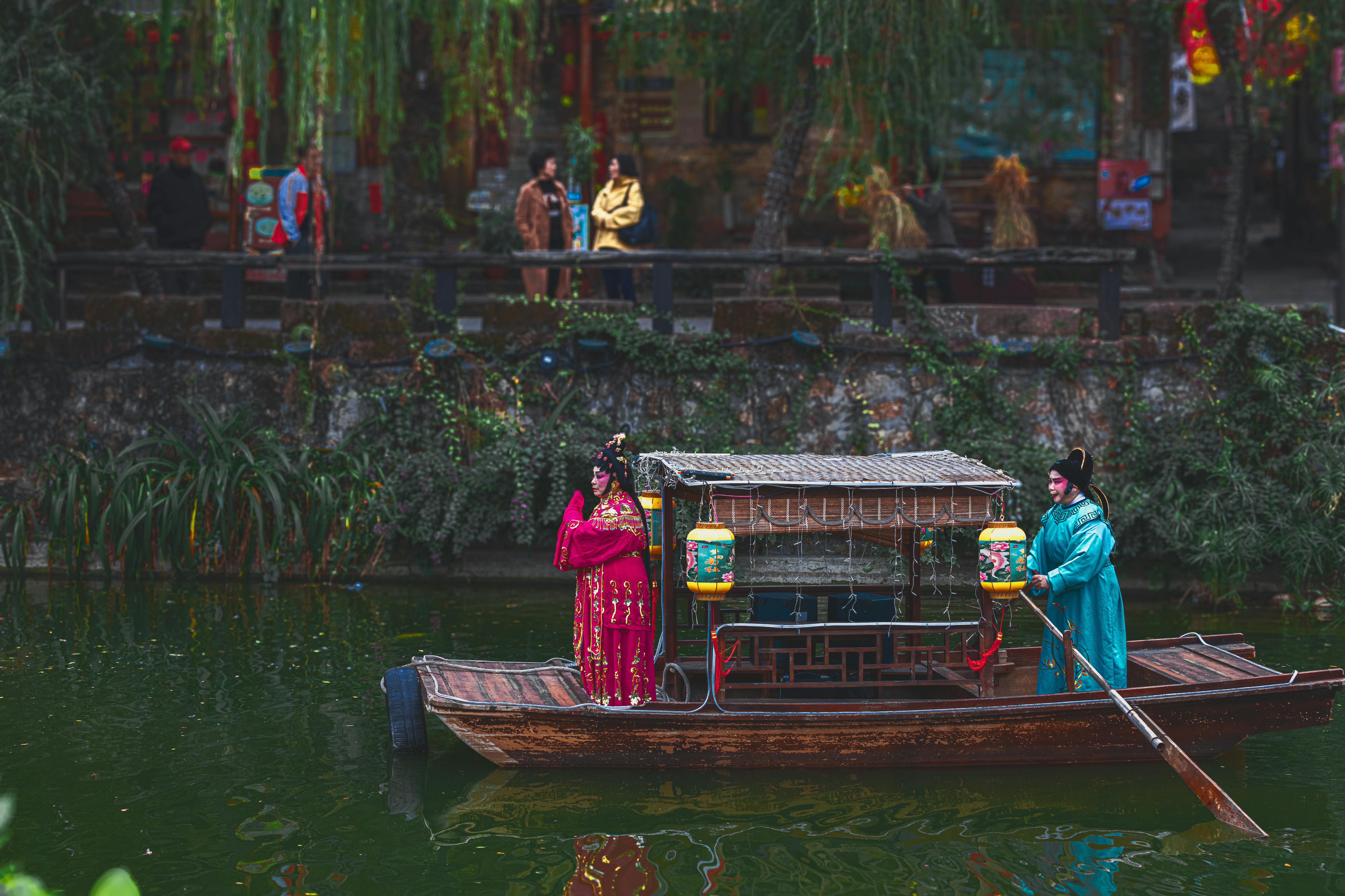 Performers in traditional costumes on a wooden boat in an ancient canal setting, attracting tourists.