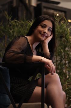 Portrait of a smiling woman in black clothes, sitting indoors with hand on cheek.