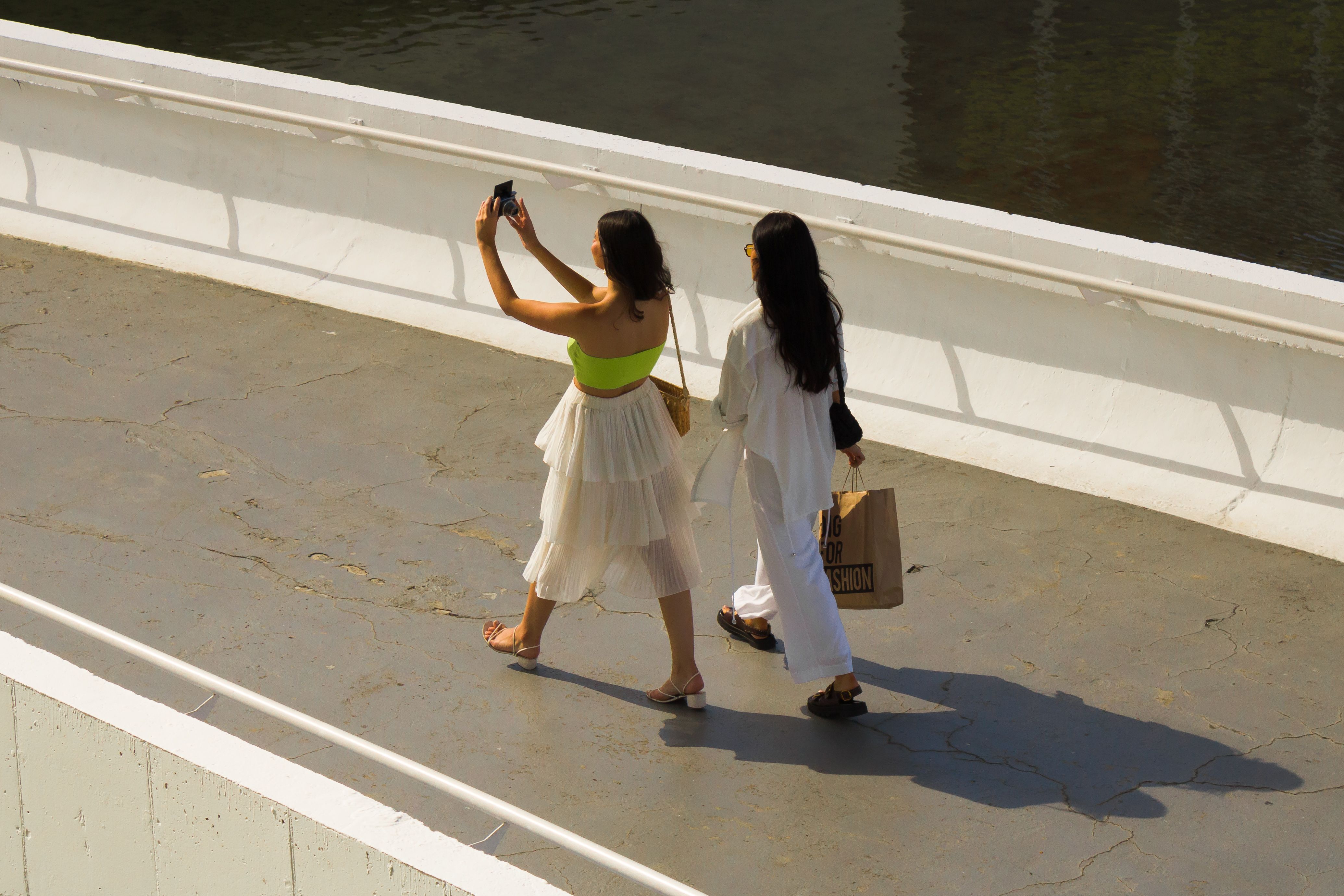 Women taking photos while walking on an urban footbridge, capturing memories.