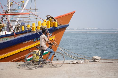 Free Man cycling on a seaside promenade with a vibrant fishing boat in the background. Stock Photo