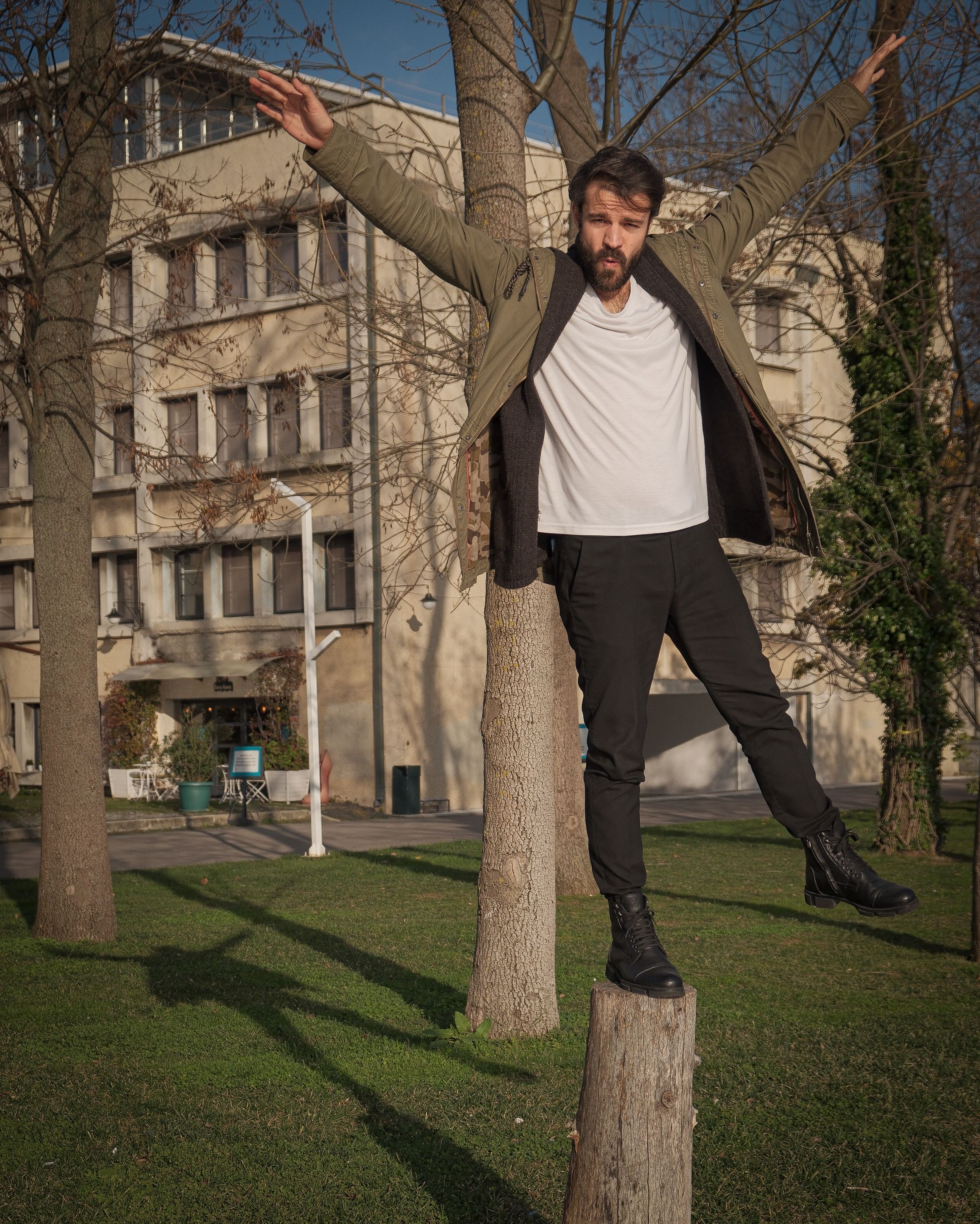 Man in stylish urban outfit balancing on a tree stump in a park.