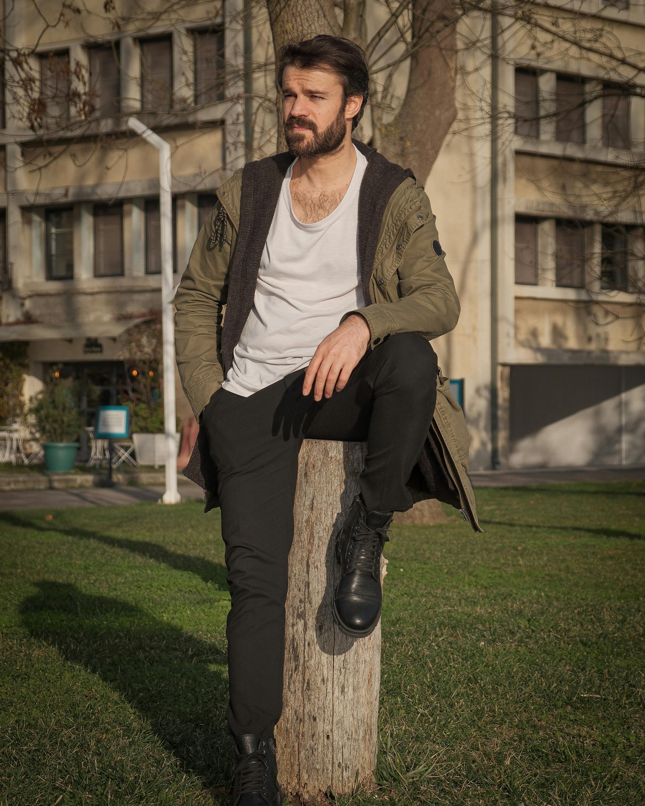 Man in fashionable attire leaning on a tree stump in an urban park setting.