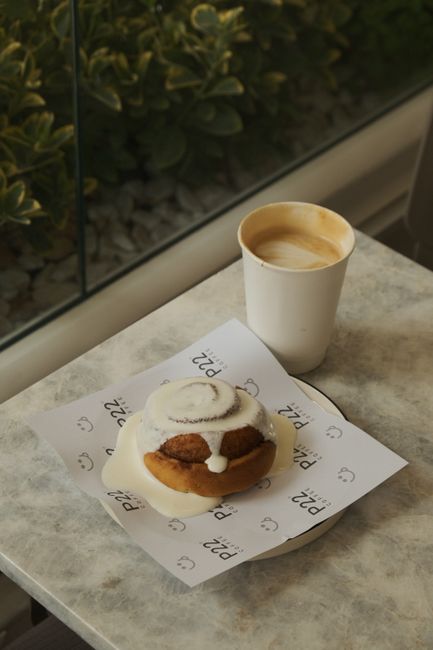 Cinnamon roll with icing and cappuccino in a paper cup on a marble table inside a cozy cafe.