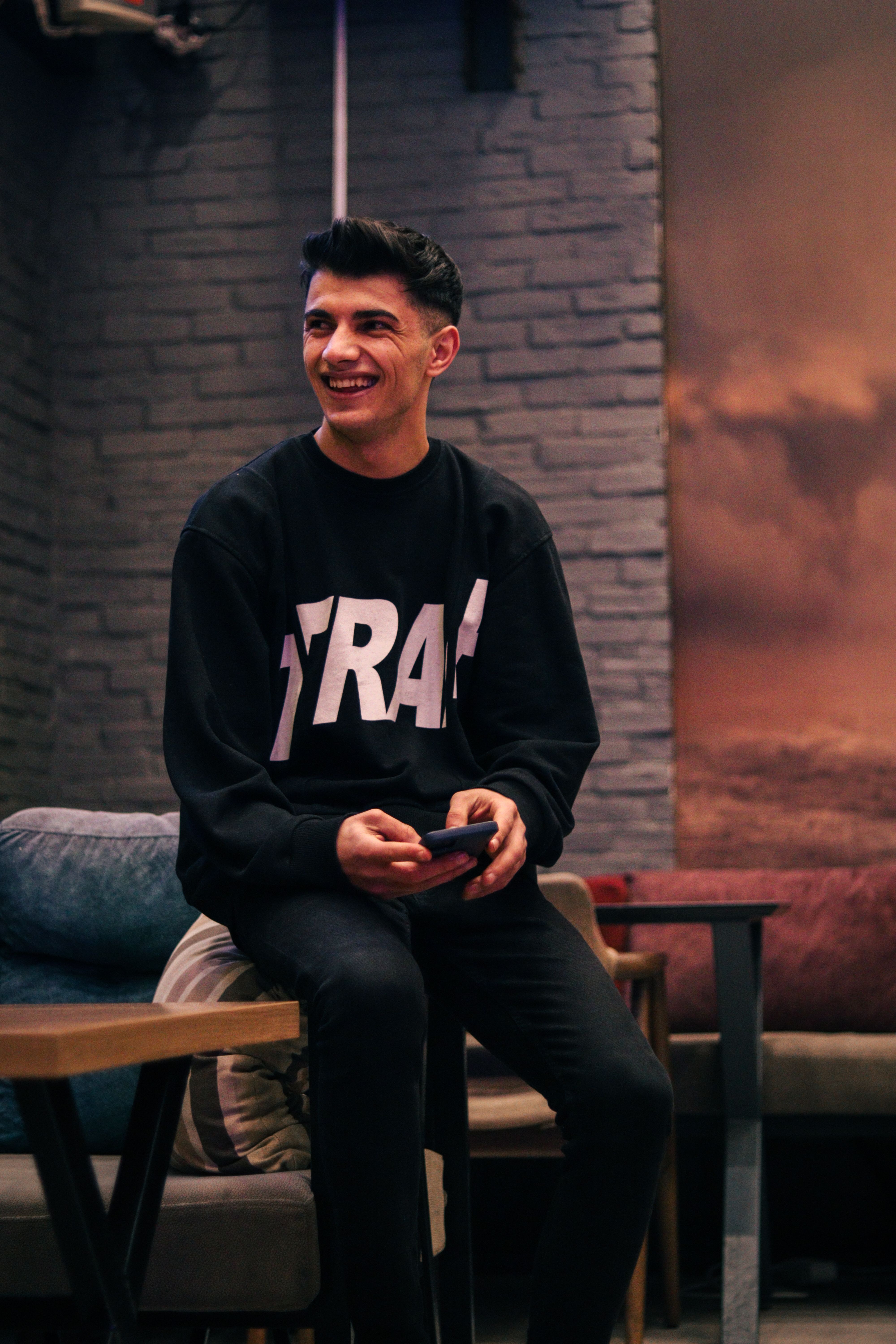 Smiling young man seated indoors holding a smartphone, wearing a trendy sweatshirt.