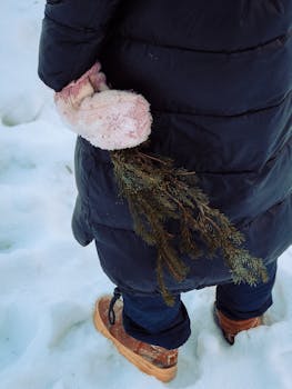 A person in a black jacket holds an evergreen branch, standing in the snow on a winter day.