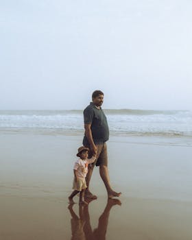 A father and son bond while walking on a sandy beach, waves crashing gently nearby.