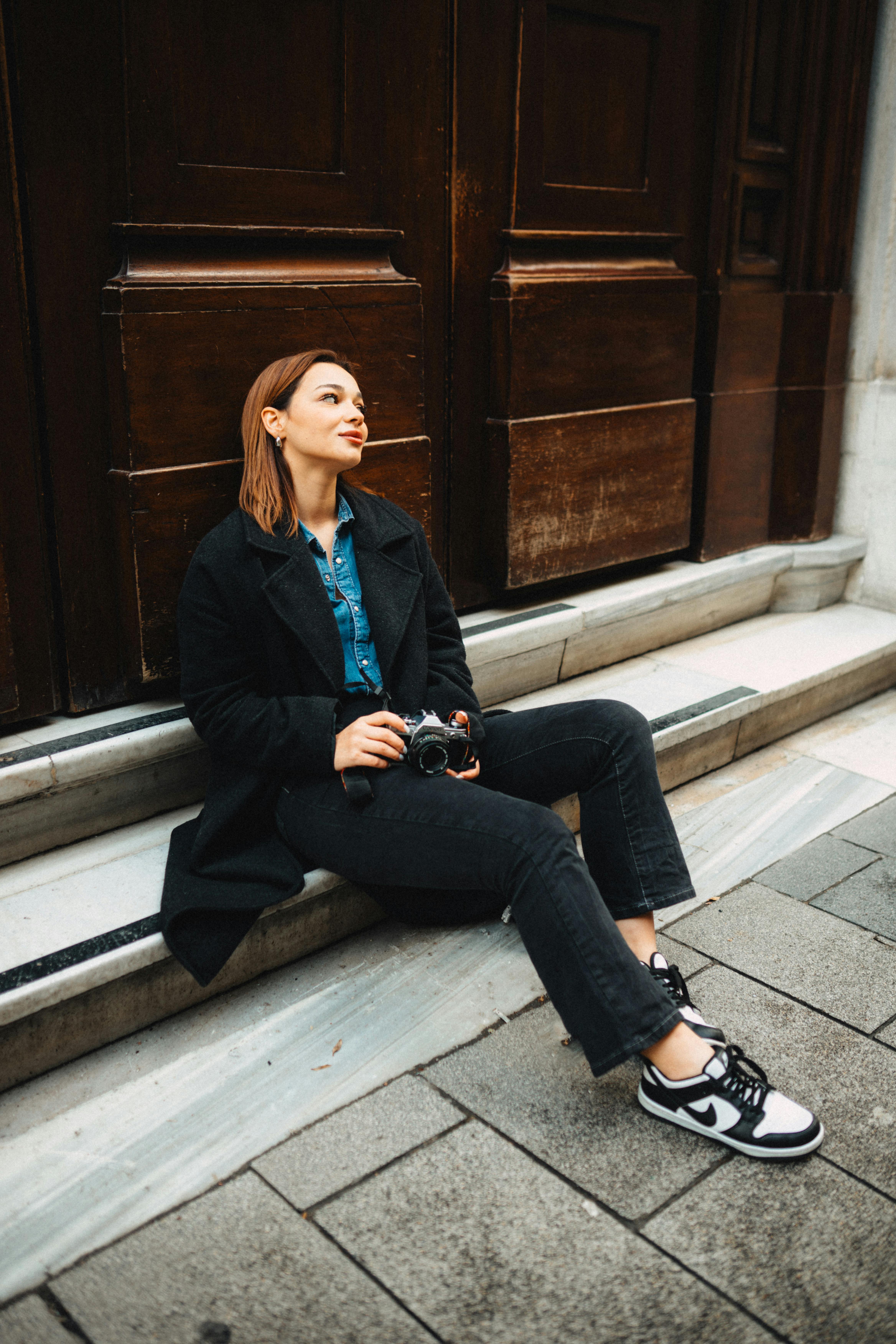 Stylish woman sitting on urban steps holding a camera, exuding modern fashion vibes.