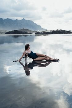 A woman in a swimsuit lounging on a calm beach shore with mountains in the background.