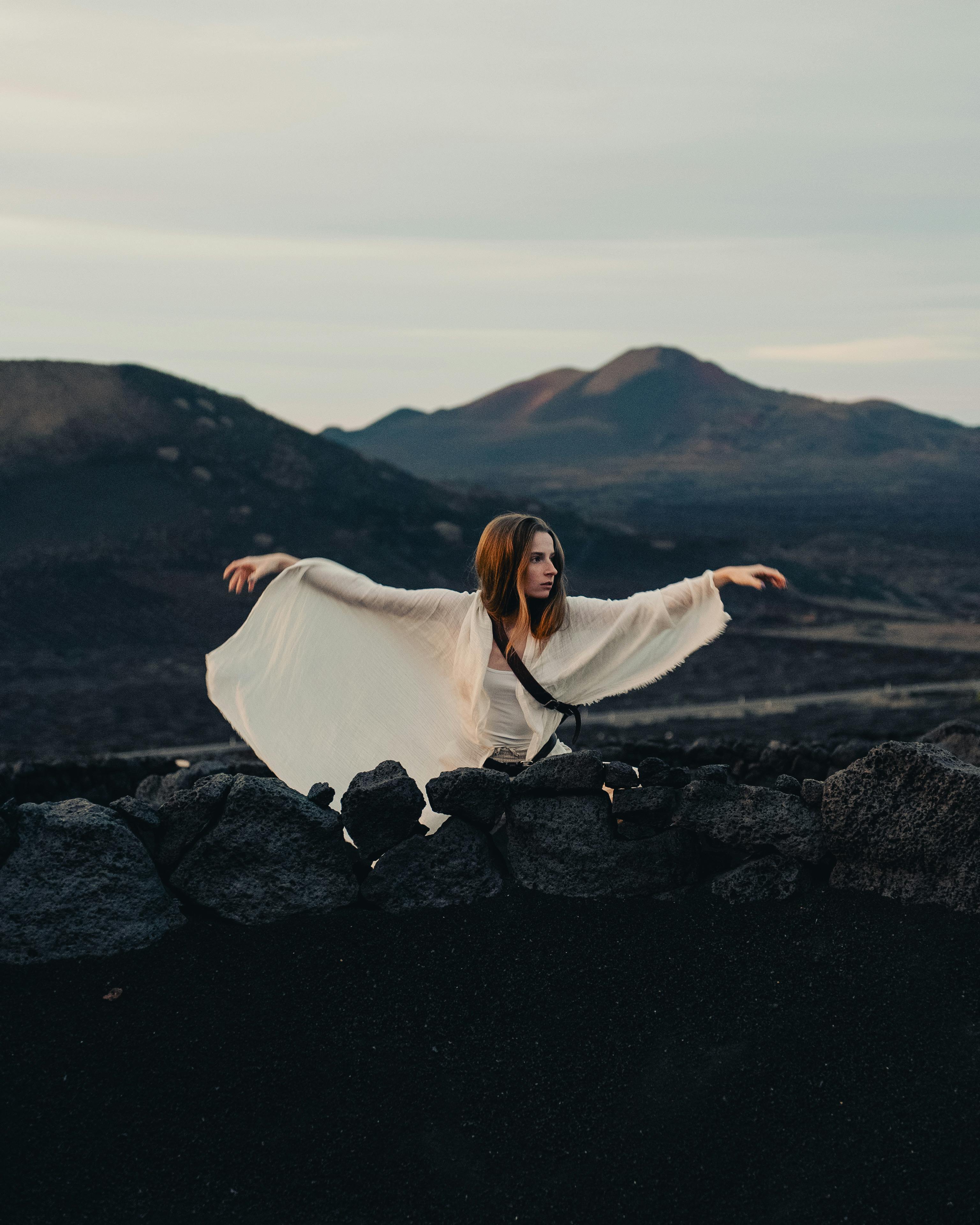 A woman in a flowing dress poses on a stone wall amidst volcanic landscape during twilight.