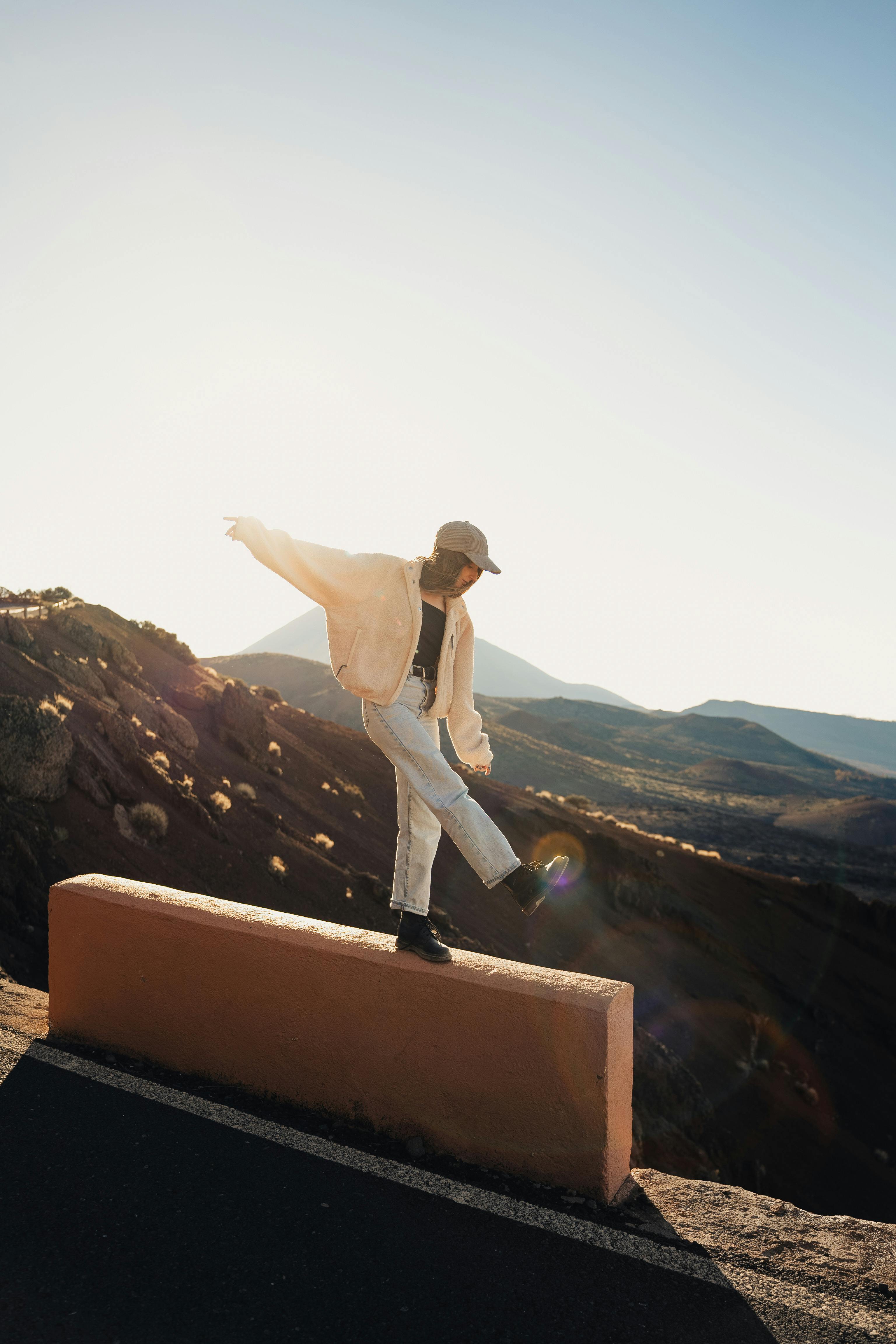 A young woman in casual attire balances on a wall with a scenic landscape backdrop during sunset.