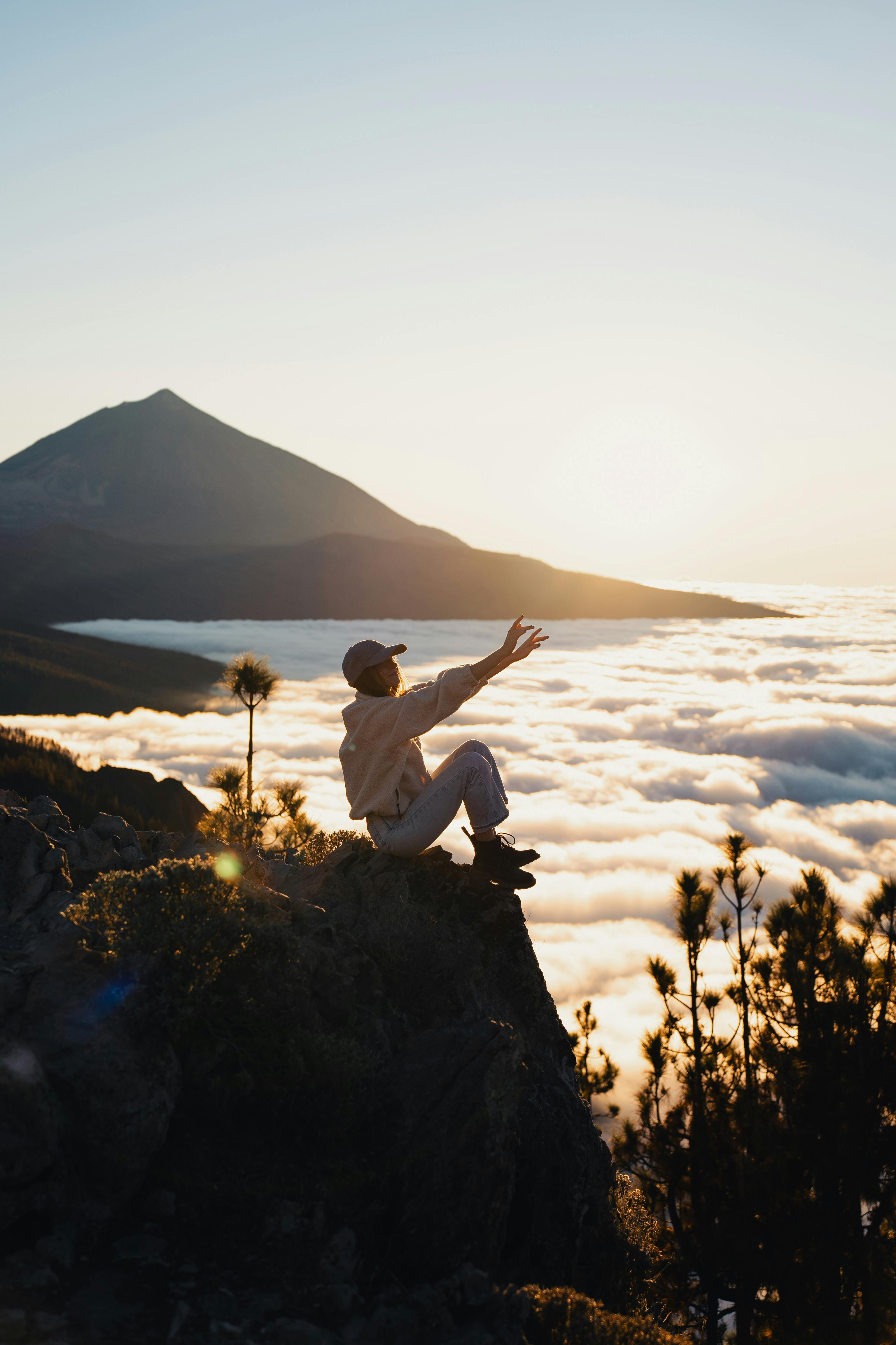 A woman sits on a mountain edge, enveloped by clouds at sunset, arms raised in serenity.