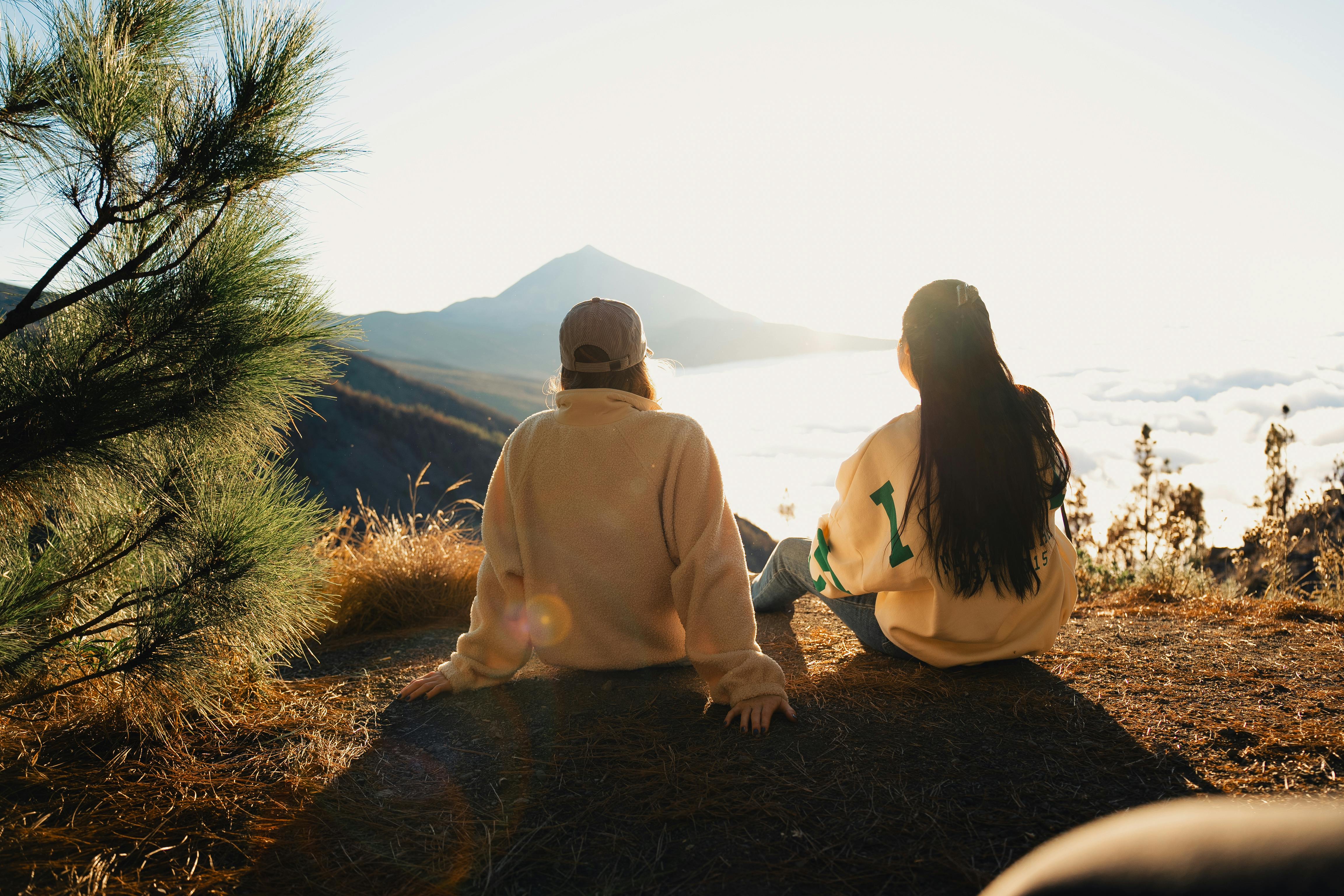 Two people sitting on a hilltop, enjoying a sunset view overlooking scenic mountains and clouds.