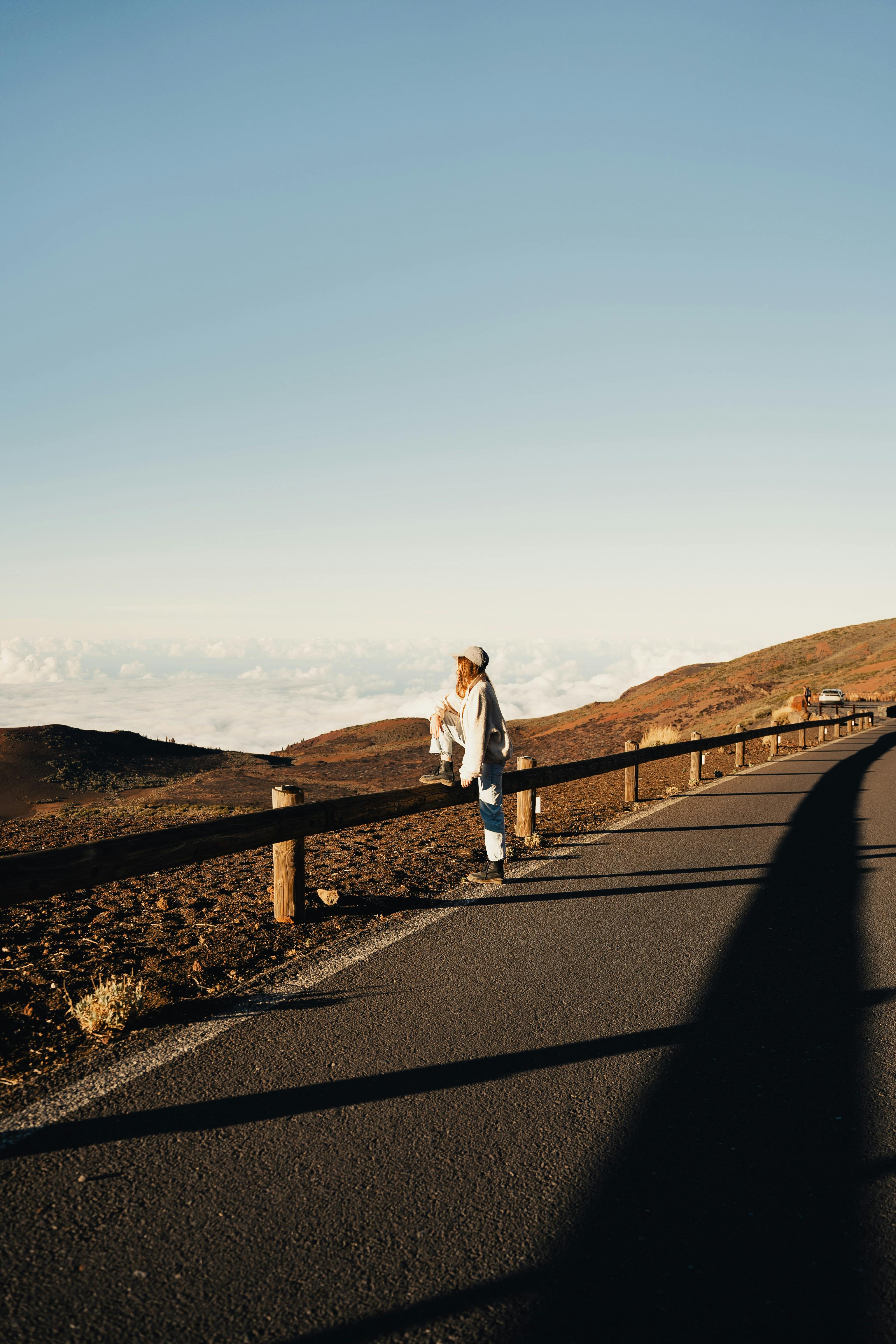 A woman in casual attire stands by a road, enjoying the vast mountain view.