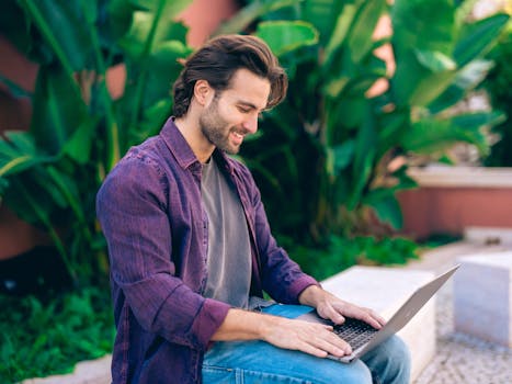 A smiling man in Lisbon using a laptop outdoors, surrounded by lush greenery. Perfect for lifestyle and remote work themes.