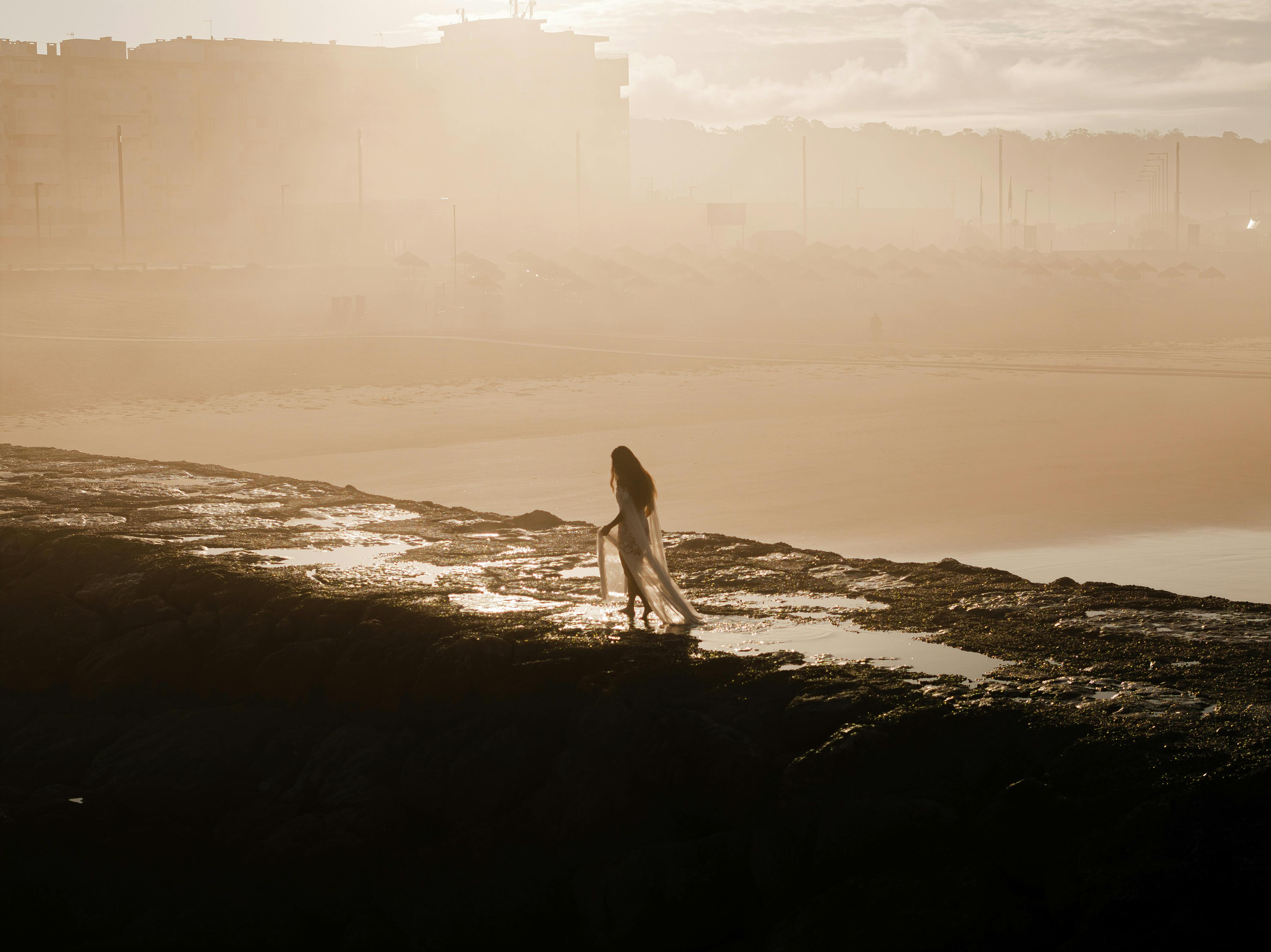 A woman in a white dress strolls along a rocky coastline at sunset, Lisbon, Portugal.