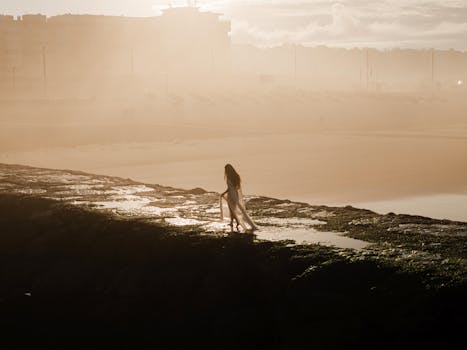 A woman in a white dress strolls along a rocky coastline at sunset, Lisbon, Portugal.