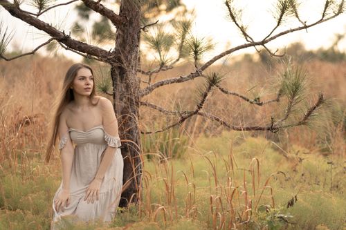 Free Woman in off-shoulder dress kneeling by tree in serene nature setting. Stock Photo