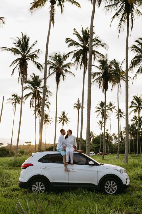 Free A couple enjoys a romantic moment on an SUV amidst tropical palm trees during sunset. Stock Photo