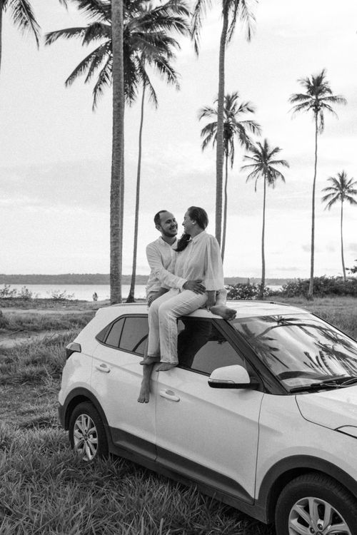 Free A couple sharing a moment on a car roof amid tropical palm trees by the sea. Stock Photo