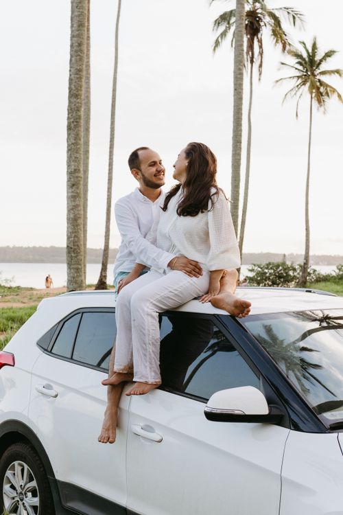 Free Couple in white outfits sitting on a car roof near palm trees by the beach, enjoying a sunny day. Stock Photo