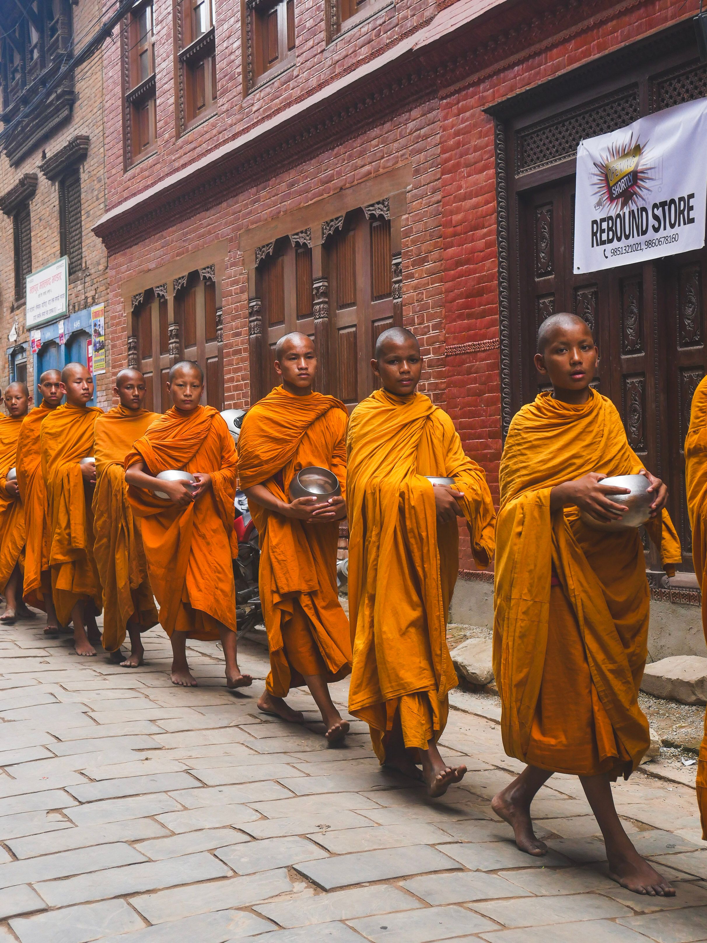 A line of young Buddhist monks walking on urban pavements in vibrant yellow robes, performing a traditional ritual.
