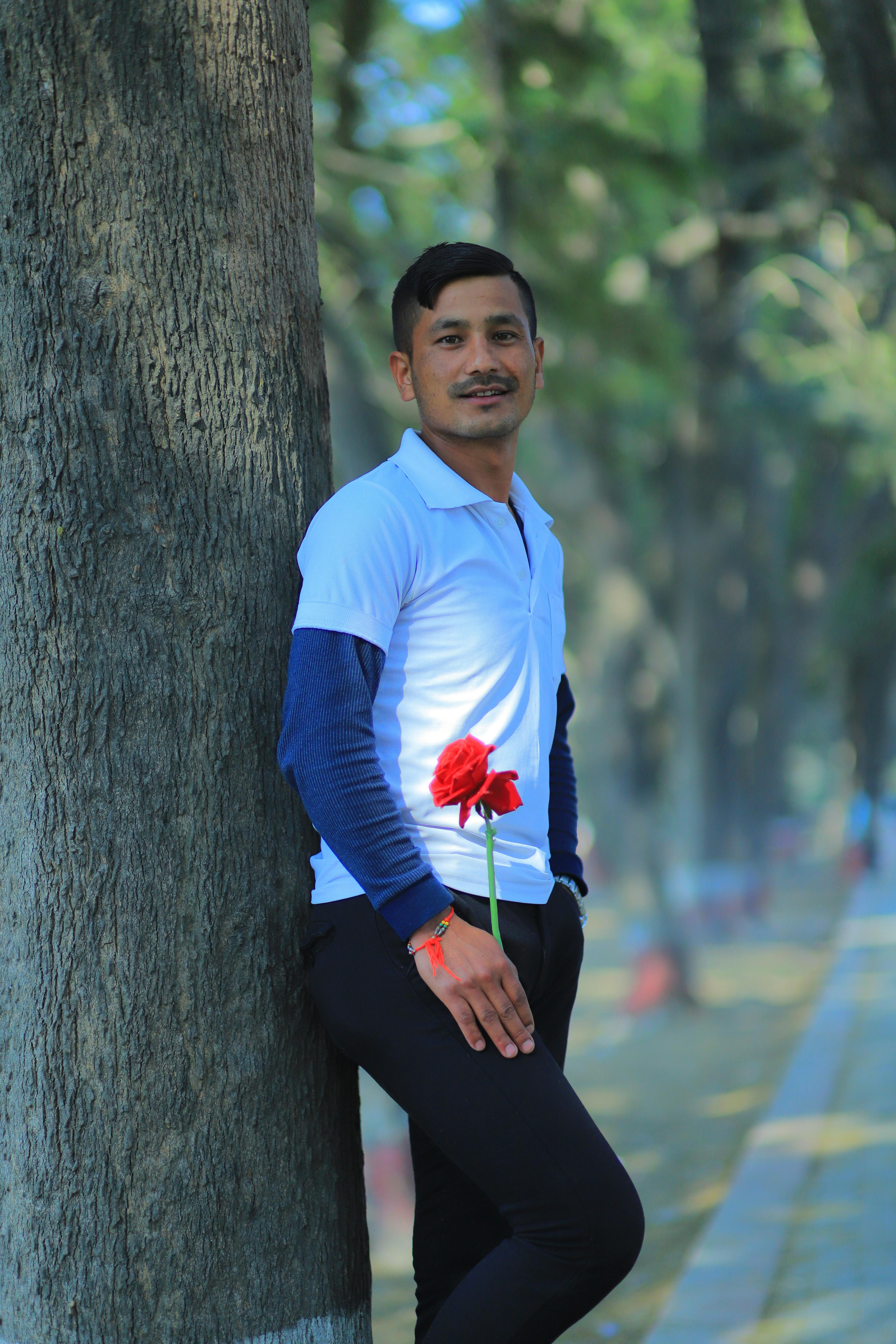A man in a white shirt smiling and holding a red flower, leaning against a tree outdoors.