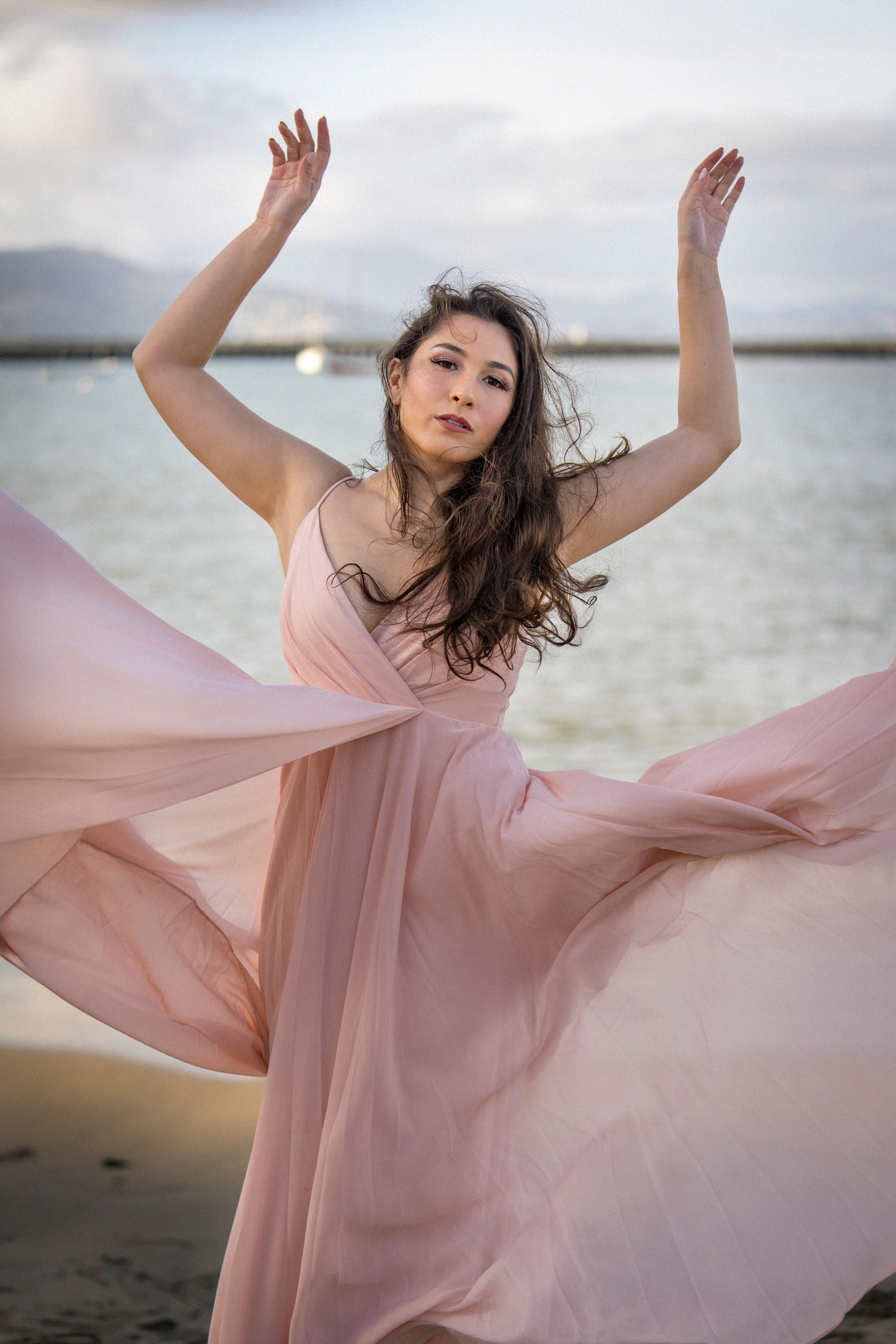 A graceful woman in a pink dress poses dramatically on a beach in San Francisco, evoking a sense of beauty and freedom.
