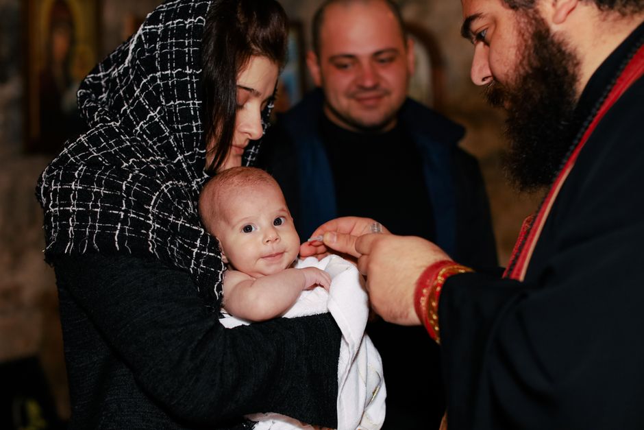 A traditional Orthodox baptism featuring a priest, baby, and family in an intimate ceremonial setting.