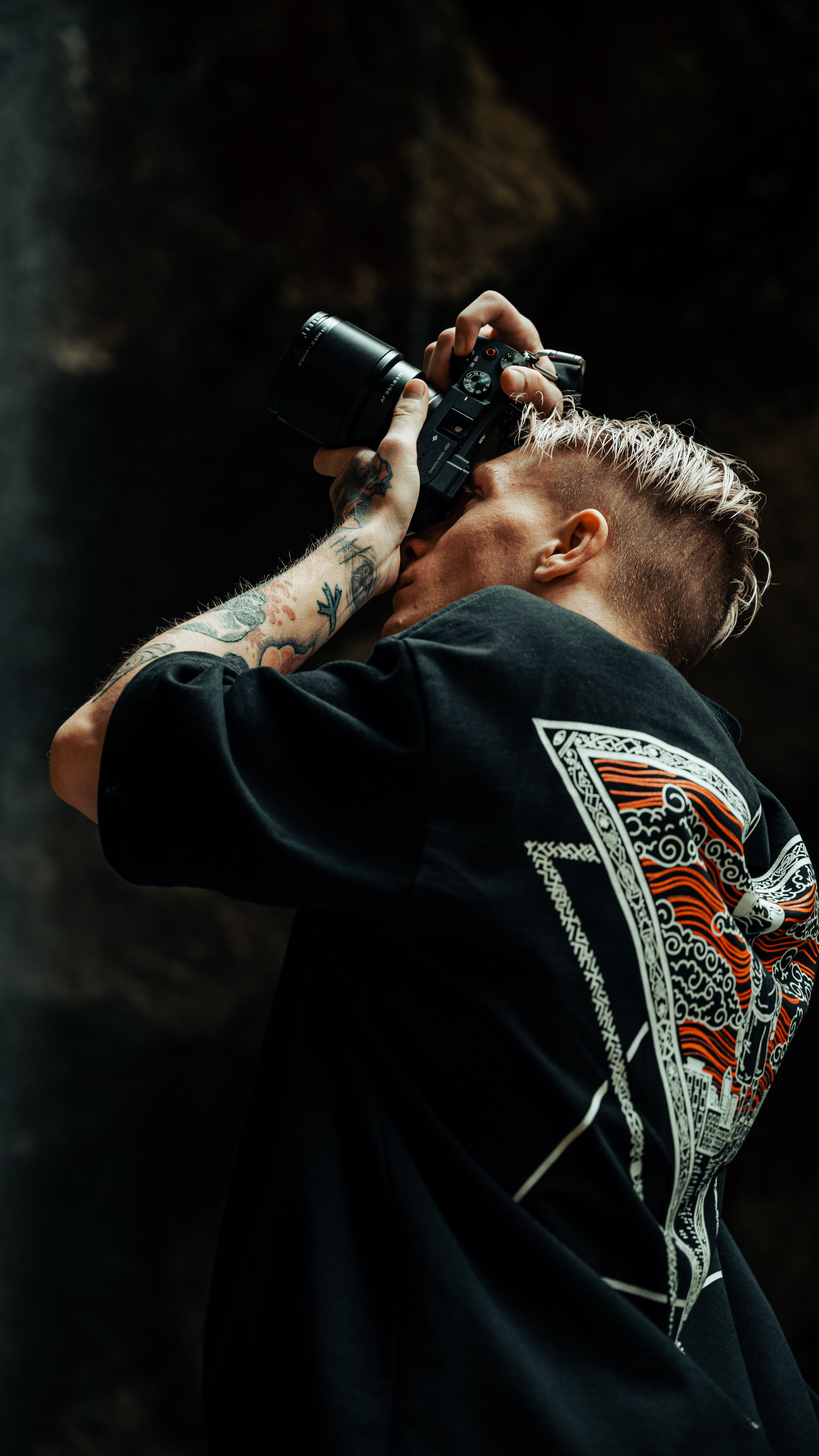 A man with tattoos and short hair uses a camera outdoors, capturing images in a natural setting.