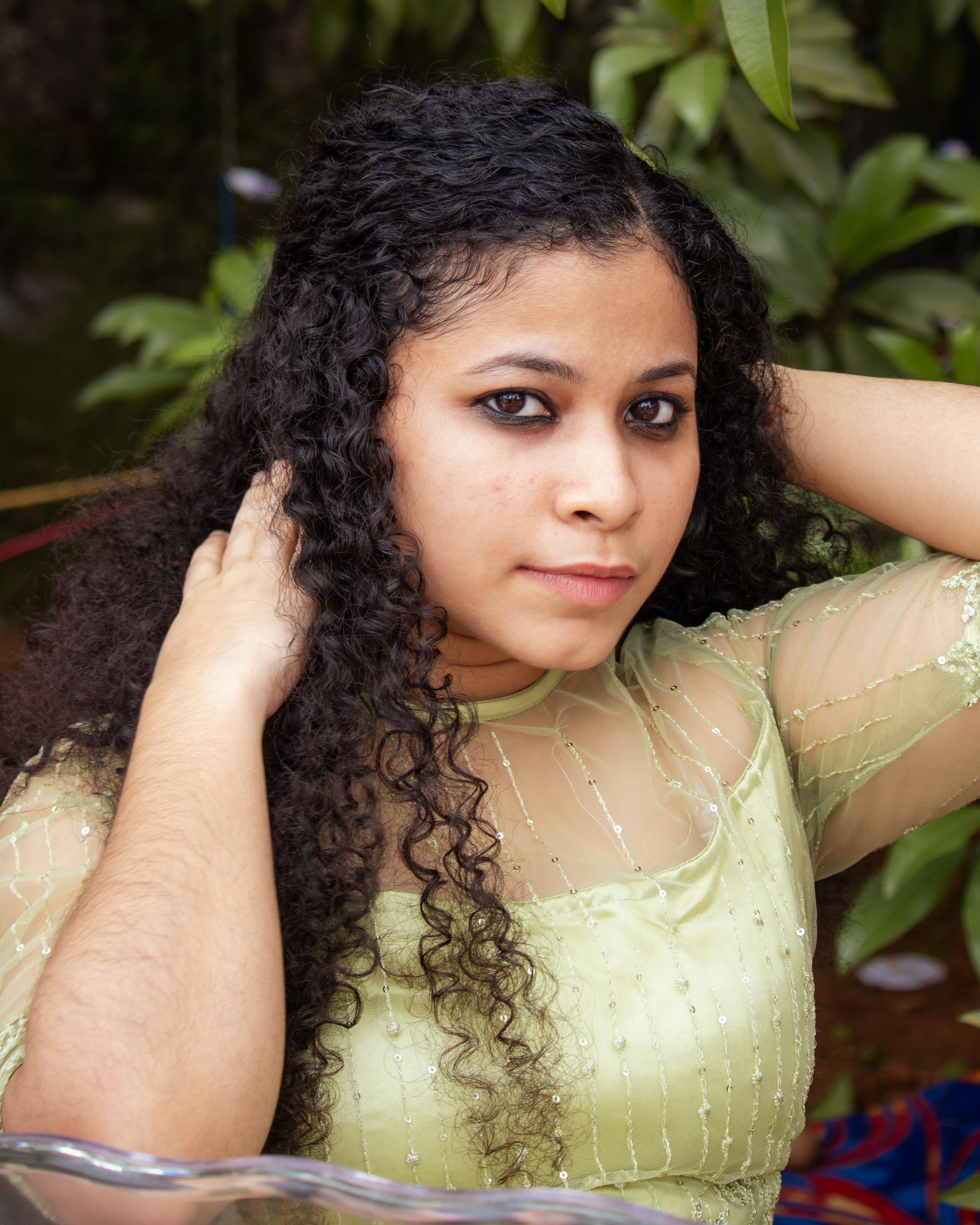 A young woman with curly hair poses outdoors in an elegant dress, hands in her hair, creating a serene look.