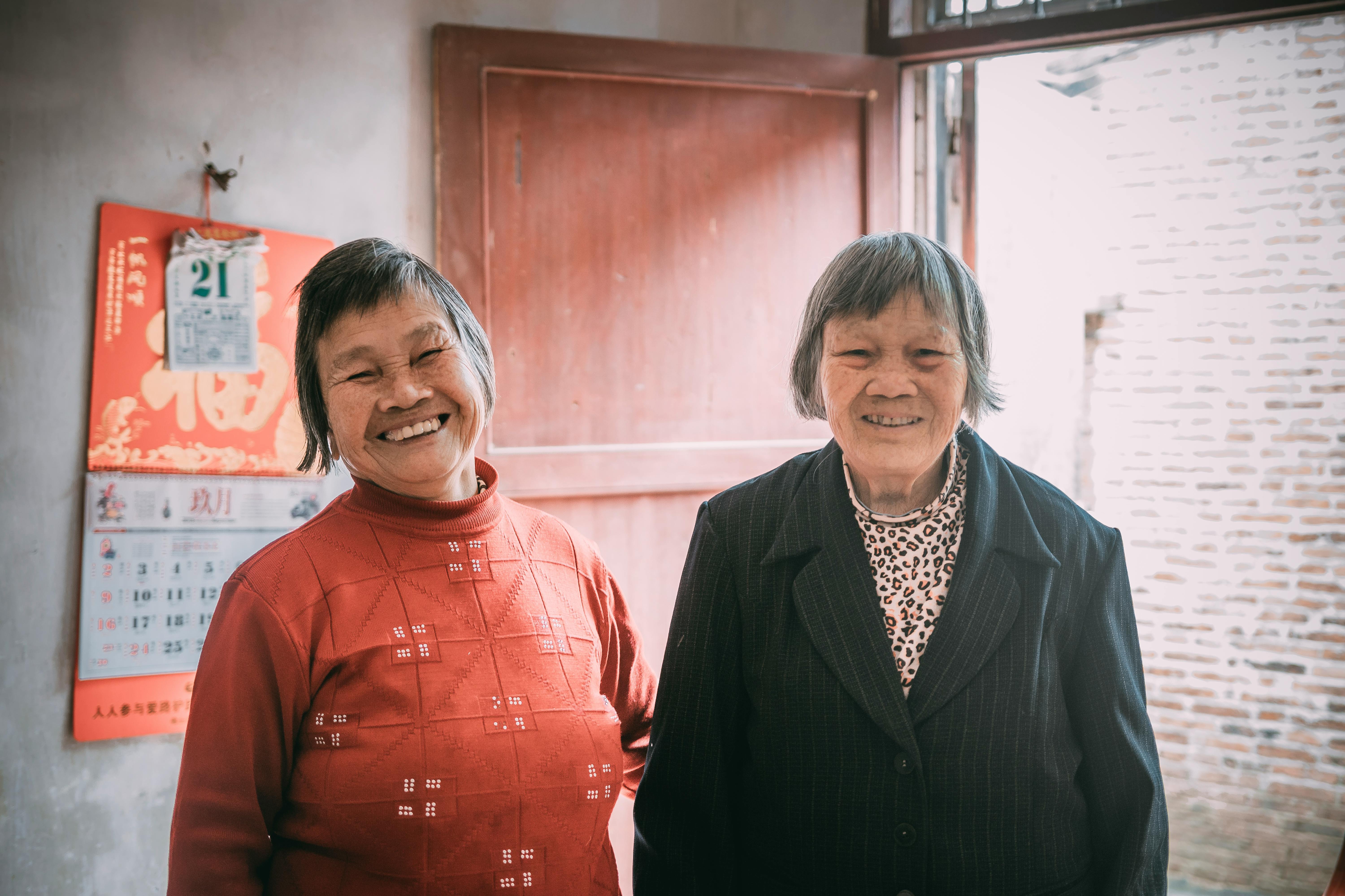 Two elderly Asian women smiling indoors, showcasing friendship and warmth.