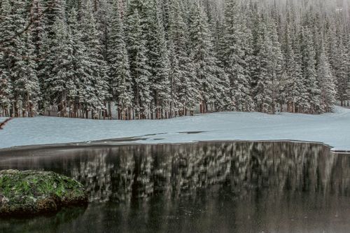 Free A tranquil winter scene of a snowy forest reflected in a partially frozen lake, capturing nature's serene beauty. Stock Photo