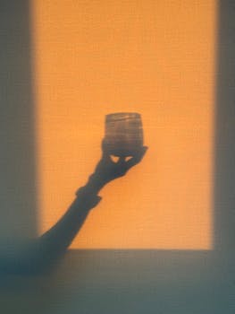 Artistic shadow of a hand holding a glass, captured in warm sunset lighting on a textured background.