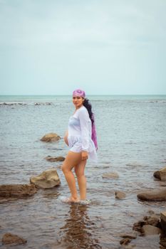 A pregnant woman poses confidently on a rocky seashore with a serene ocean backdrop.