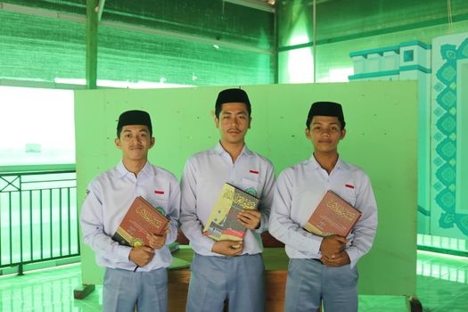 Three male students in school uniforms holding textbooks, pose indoors against a green backdrop. - Fotografer Sidoarjo