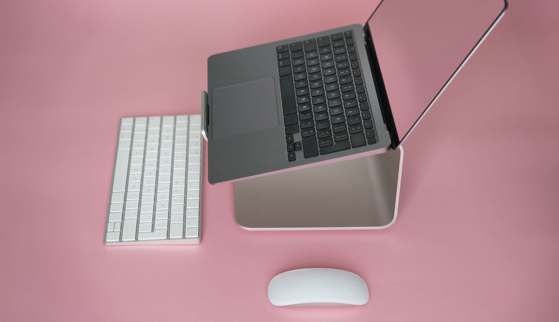 Elegant laptop setup with keyboard and mouse on pink background, showcasing sleek design and technology.