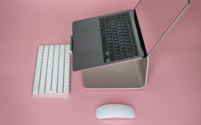 A side view of a laptop on a curved silver stand on a pink desk, with a white keyboard and mouse